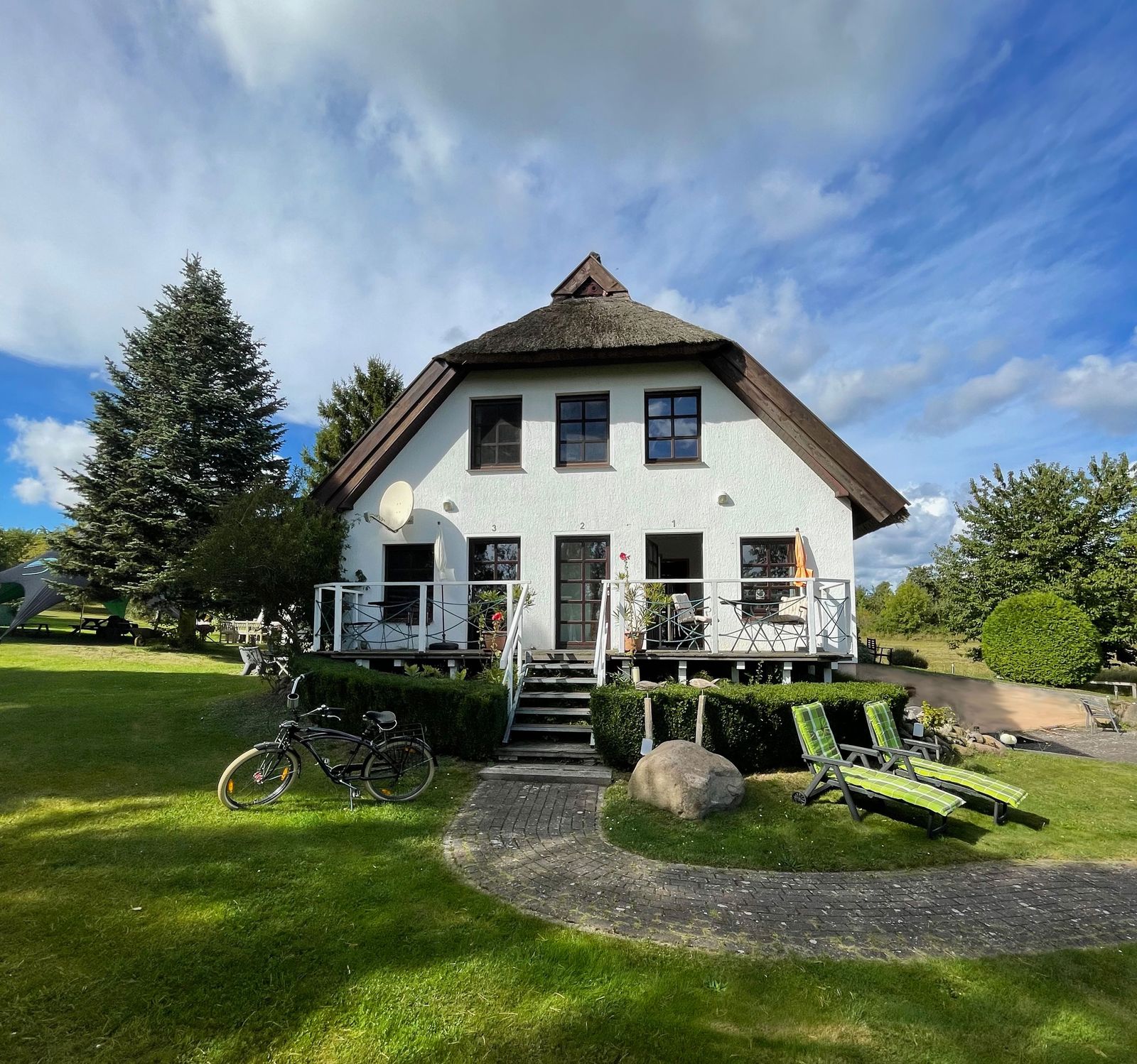 Das weiße Reetdachhaus Dat Witte Hus in Moritzdorf mit Gästeeingang, Treppe zur Terrasse, Liegestühlen und Fahrrad auf dem Rasen unter blauem Himmel