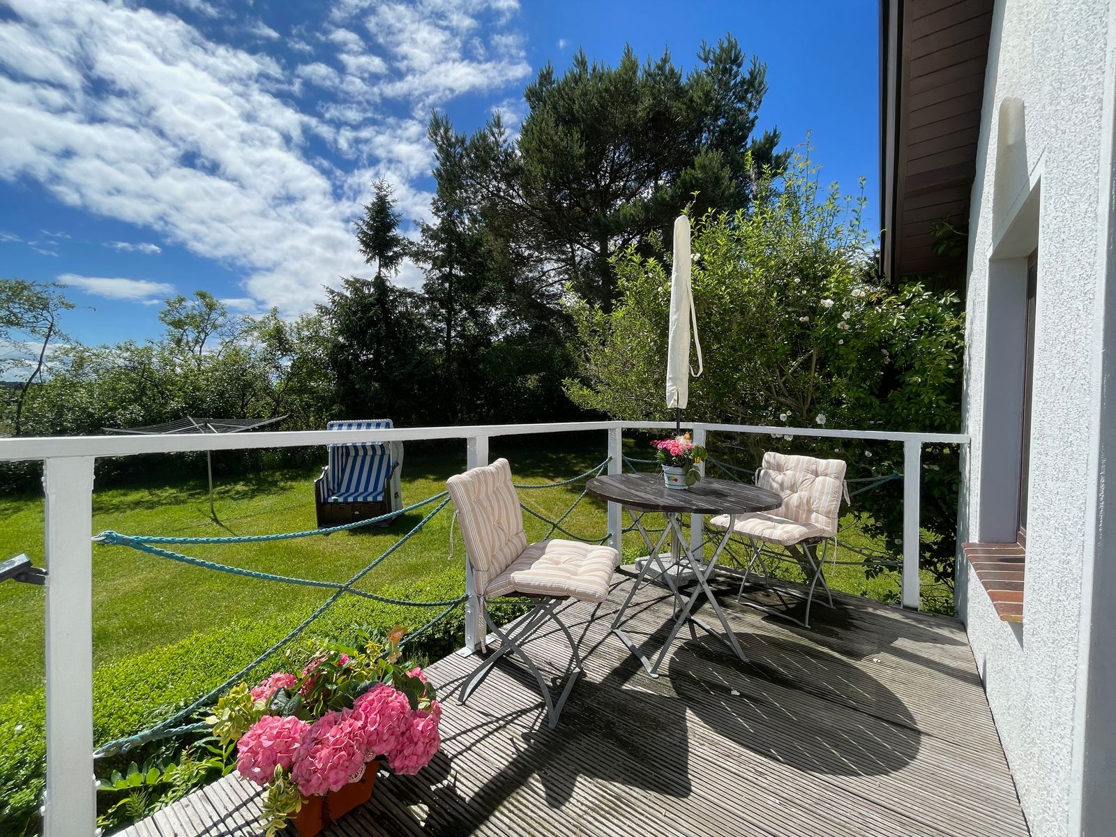 Holzterrasse am Dat Witte Hus mit Bistrotisch, zwei Stühlen und rosa Hortensien, dahinter Rasen, Strandkorb und hohe Kiefern