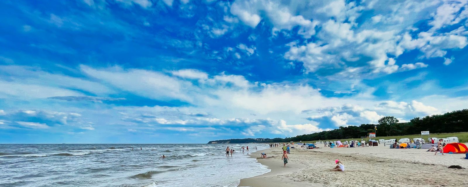 Weiter Ostseestrand an einem Sommertag mit Badegästen, bunten Sonnenschirmen und bewaldeter Küste im Hintergrund