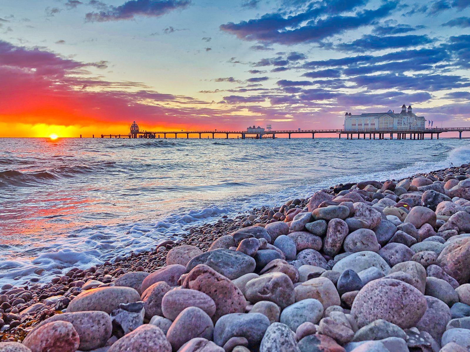 Die Seebrücke Sellin mit ihrem weißen Brückenhaus im Licht der aufgehenden Sonne, im Vordergrund rund geschliffene Steine am Strand